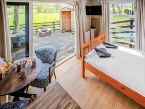 A bedroom with a bed, a table with a chair, a TV, and a view of a grassy area.