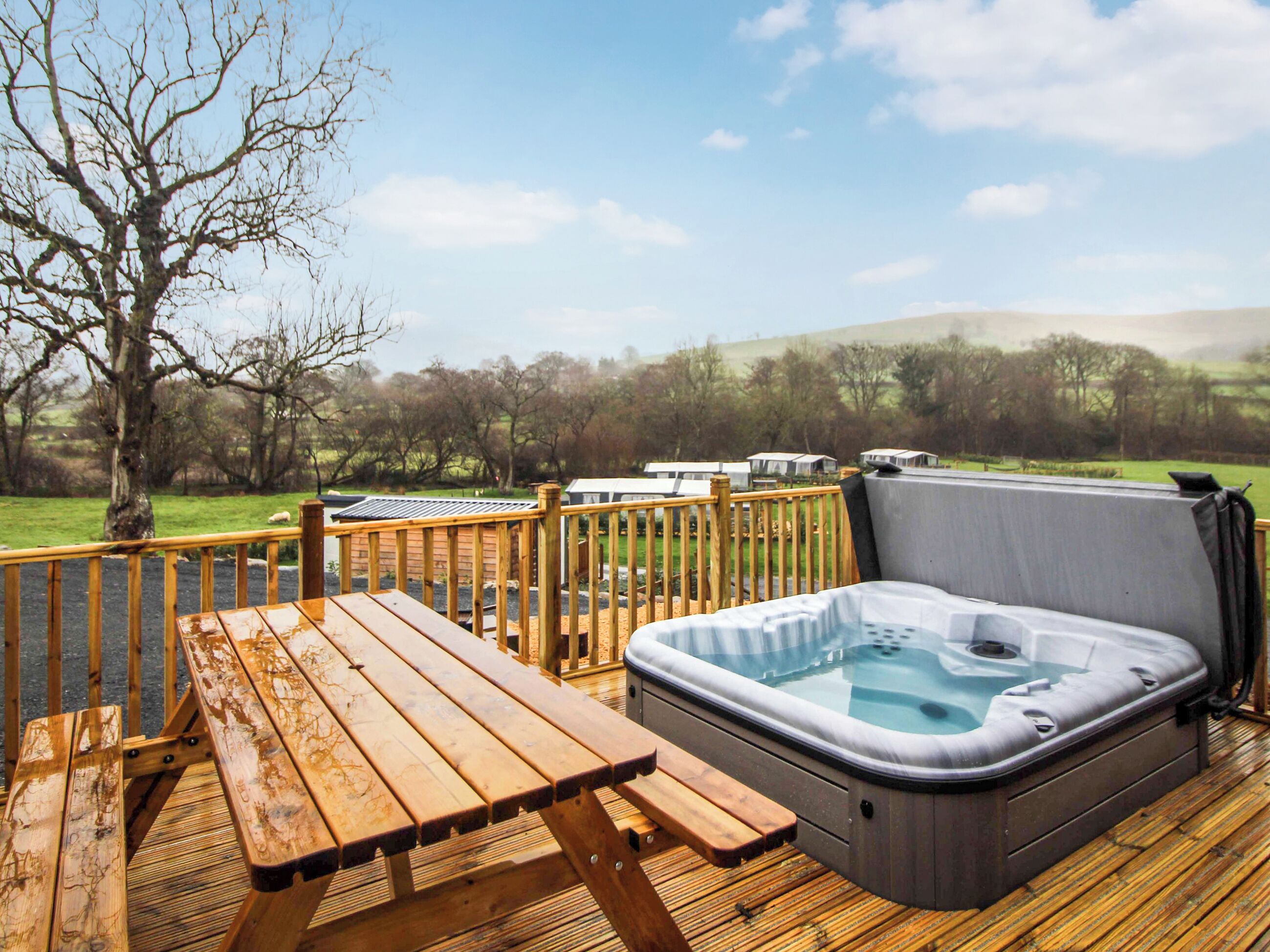 A wooden deck with a hot tub and picnic table, overlooking a grassy landscape with trees and hills.