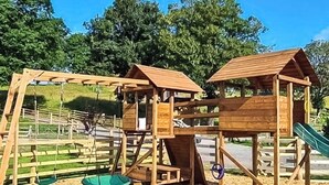 Une structure de jeux en bois, avec des balançoires et un toboggan, entourée d’arbres et d’une zone herbeuse.