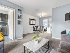 Een moderne woonkamer met een marmeren salontafel, een eethoek met een witte tafel en stoelen, een keuken met roestvrijstalen apparatuur, en een aan de muur bevestigde televisie.