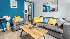 A modern living room with a blue accent wall, a grey sofa with yellow and patterned cushions, a wooden coffee table with a bowl and wine glasses, and a large mirror on the wall. - 2 bedroom accommodation in Jesmond (Jesmond)