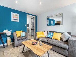 Un salon moderne avec un mur d’accent bleu, un canapé gris agrémenté de coussins jaunes et à motifs, une table basse en bois avec un saladier et des verres à vin, et un grand miroir accroché au mur.