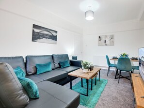 A modern living room with a grey sofa, teal cushions, a wooden coffee table, and teal chairs. A painting of a bridge is on the wall, and a dining table with chairs is in the background. - 3 bedroom accommodation in Jesmond (Jesmond)