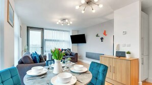 Ein modernes Wohnzimmer mit einem runden Esstisch für vier Personen, senfgelben Stühlen, einer Couch, einem an der Wand montierten Fernseher und einer Holzkommode.