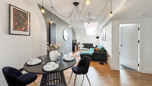 Ein moderner Essbereich mit einem runden Tisch, schwarzen Stühlen und einer Blumenvase. Im Hintergrund befinden sich eine Couch und ein Fernseher.