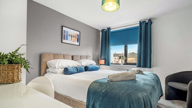 A modern bedroom with a bed, a bedside table, a lamp, a window with curtains, and a view of buildings outside. - 2 bedroom accommodation in Tyne and Wear (Tyne and Wear)
