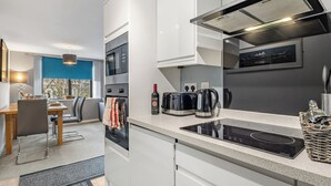 A modern kitchen with white cabinets, a built-in oven, a stove, and a microwave. There is a dining area with a table and chairs, and a window with a view of trees. - 2 bedroom accommodation in Tyne and Wear (Tyne and Wear)