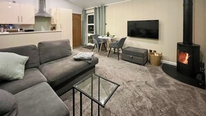 Moderni olohuone, jossa on sohva, lasinen sohvapöytä, televisio, takka sekä ruokailutila, jossa on pöytä ja tuolit.