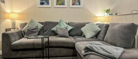A living room with a grey sectional sofa, a glass coffee table, and a lamp on a side table. There are three framed artworks on the wall.