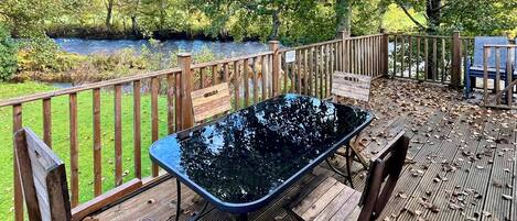 Una terraza de madera con mesa y sillas de vidrio, con vista a un río y rodeada de árboles.