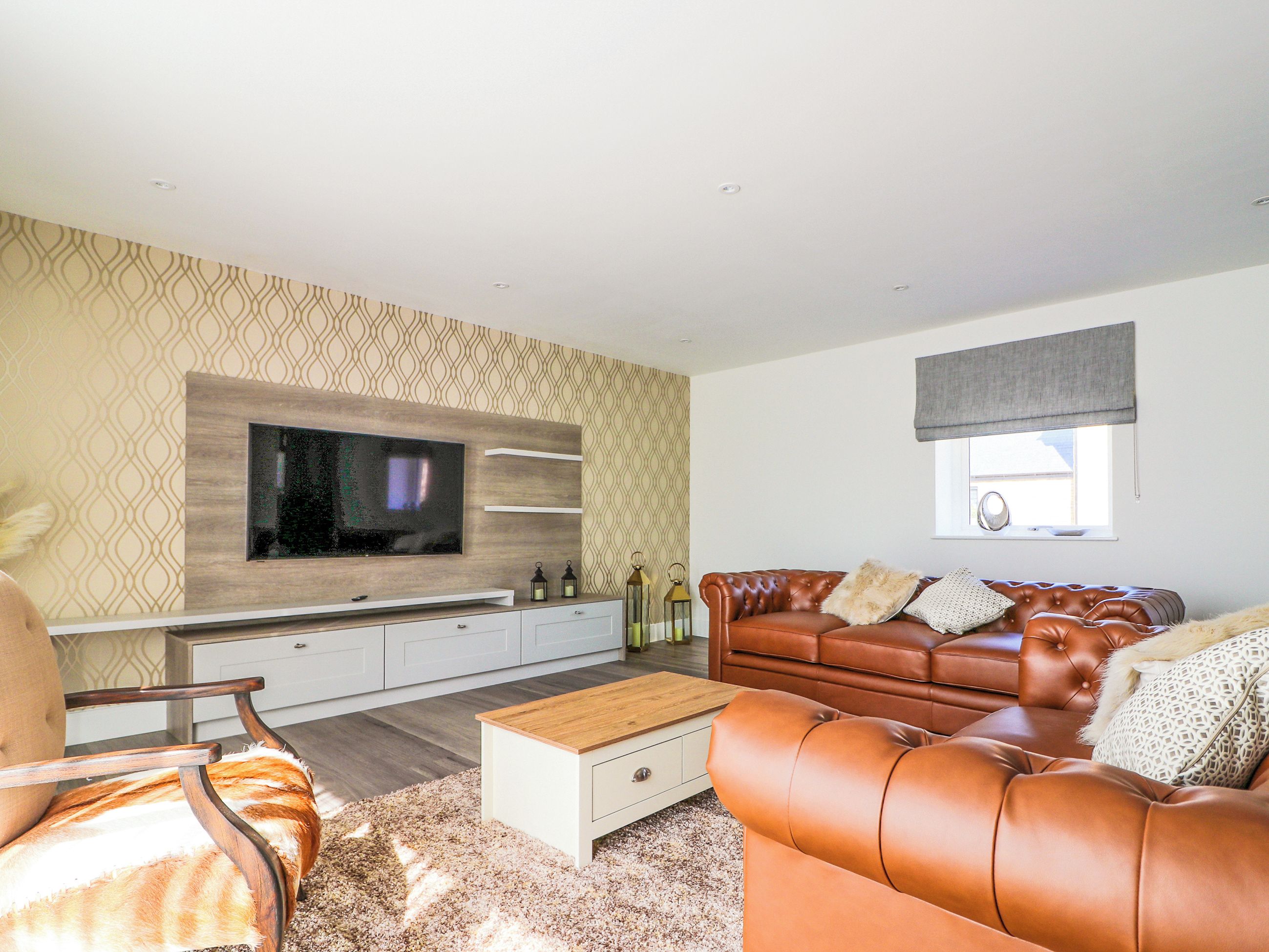 A modern living room with a flat-screen TV mounted on a wooden panel, a leather sofa, a wooden coffee table, and patterned wallpaper.