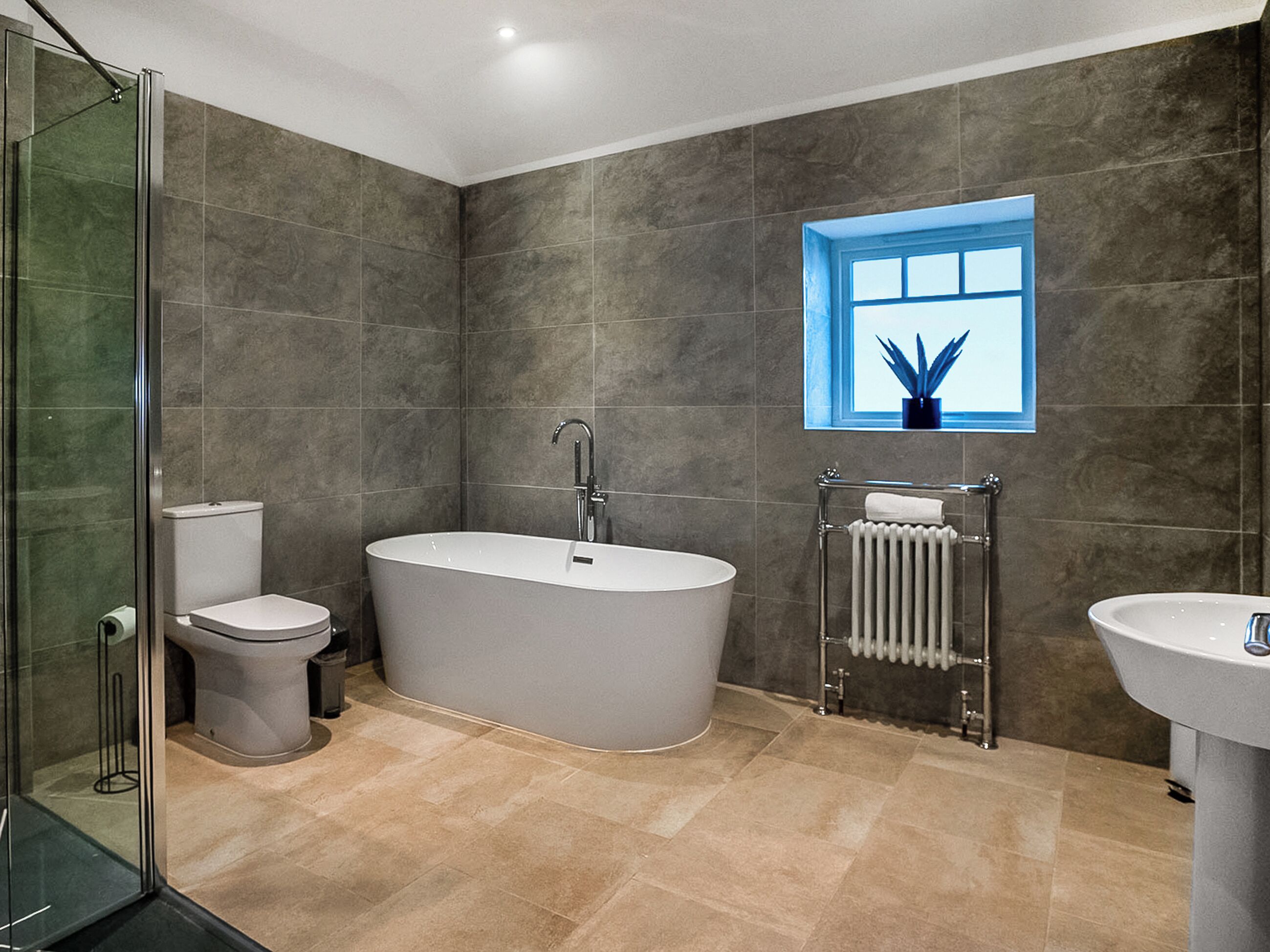 A modern bathroom with a freestanding bathtub, a toilet, a radiator, and a window with a plant.