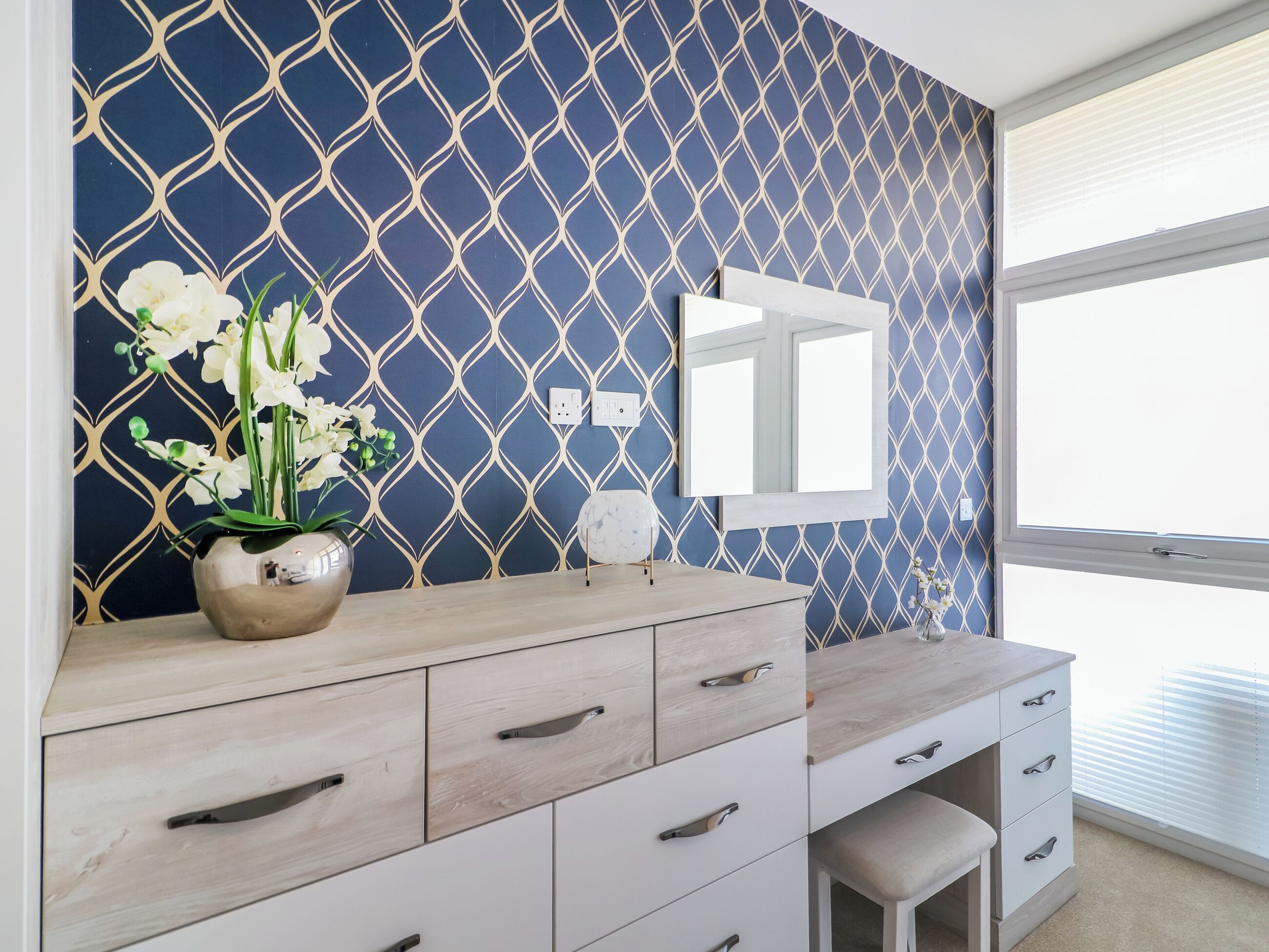 Une salle de bain avec un mur à motifs bleus, un miroir, un meuble-lavabo avec des tiroirs et une fenêtre avec des stores.