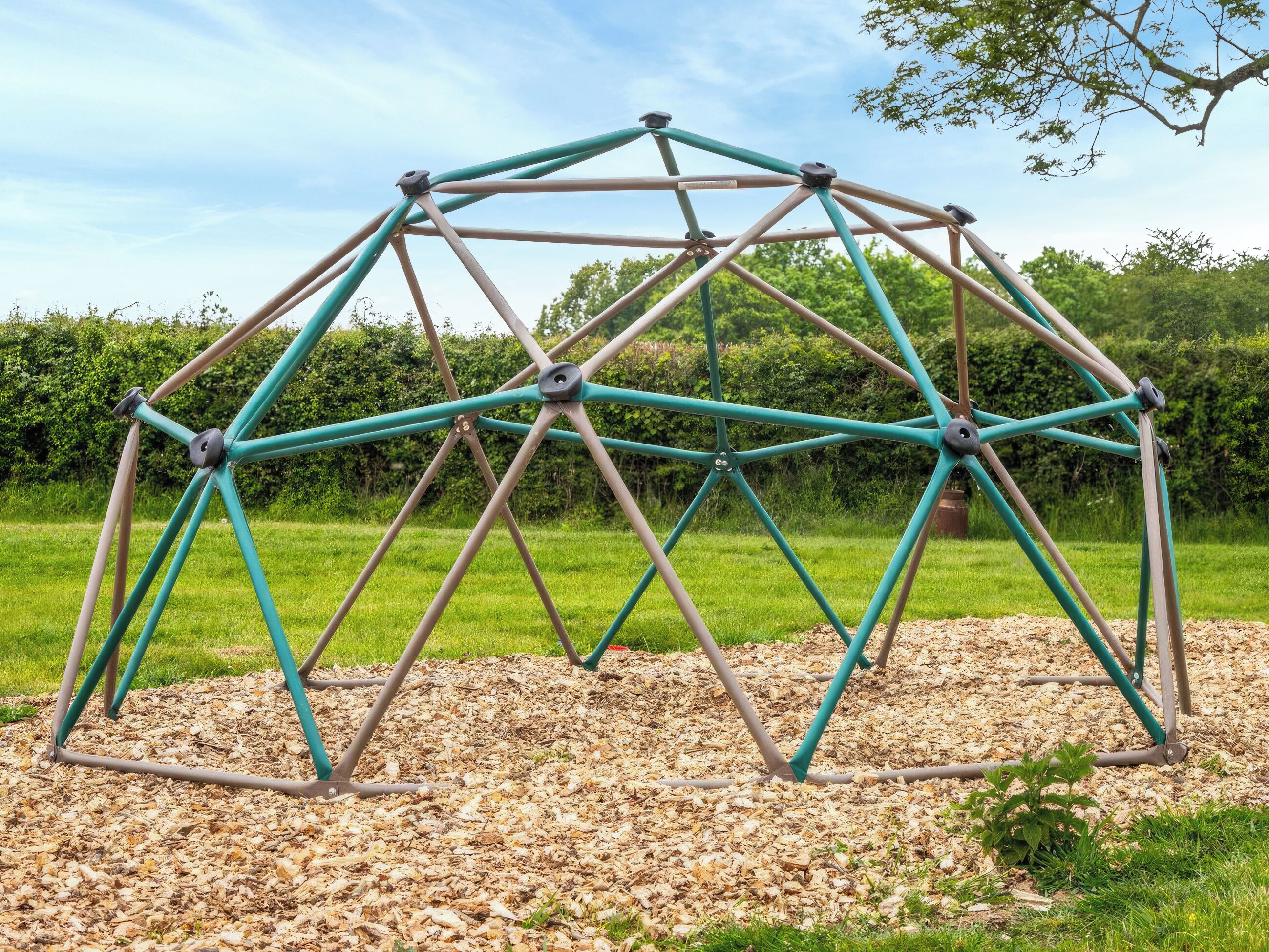 A geodesic dome structure made of metal rods and nodes, set on a bed of wood chips in a grassy outdoor area.