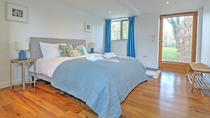 A bedroom with a large bed, wooden flooring, a window with curtains, and a view of greenery outside. - The Cart Shed - UKC8980 (Romsey)