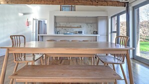 A spacious dining area with a large wooden table and chairs, a kitchen with modern appliances, and large windows offering a view of the outdoors. - The Cart Shed - UKC8980 (Romsey)