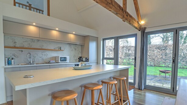 A modern kitchen with a large island, bar stools, and a view of the garden through large windows. - The Cart Shed - UKC8980 (Romsey)