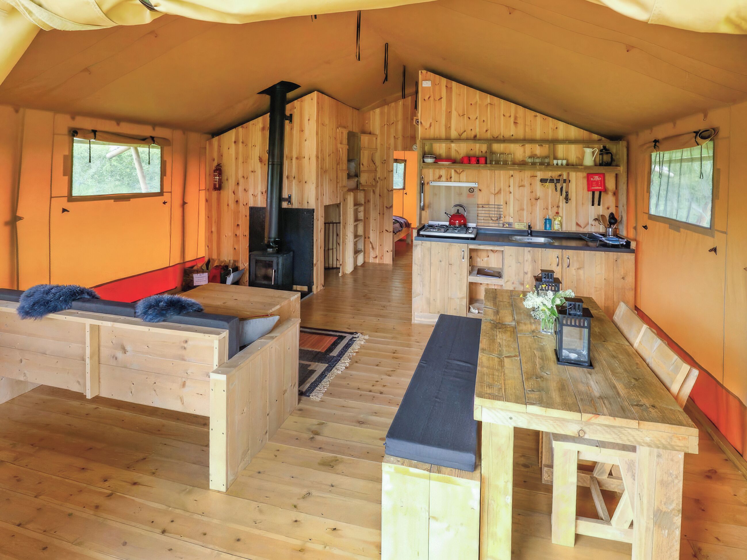 L’intérieur chaleureux d’un chalet, avec un décor en bois, une cheminée centrale, un coin repas et une cuisine dotée de meubles en bois et d’un poêle.