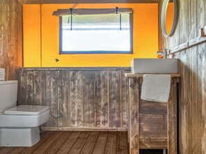 Een badkamer met houten vloer, toilet, houten wastafel met spiegel en raam met gordijn.