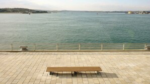 Un banc sur une promenade dallée, donnant sur une étendue d’eau calme, avec, au loin, des terres et des bâtiments.