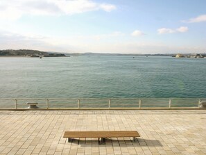 A bench on a tiled promenade overlooking a calm body of water with distant land and buildings.
