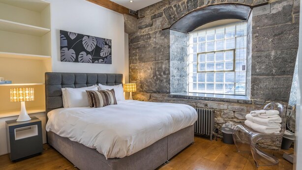 Une chambre à coucher avec un lit, deux tables de chevet avec des lampes, un mur en pierre, une fenêtre avec des stores et une chaise transparente.