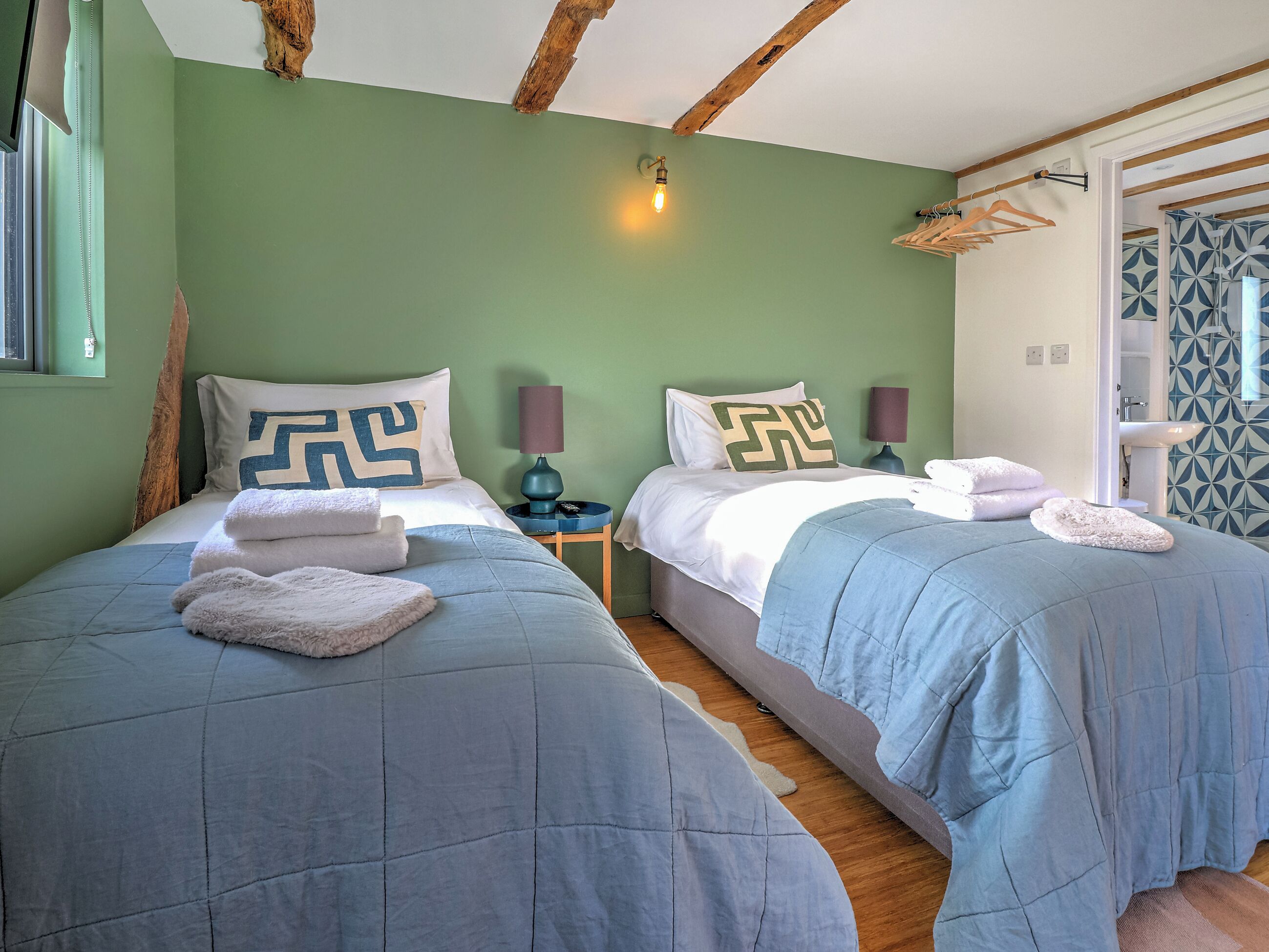 A room with two beds, each with a blue quilted bedspread and matching pillows. There are two bedside lamps, a wooden headboard, and a window with a curtain.
