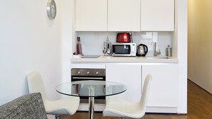 Una cocina compacta con armarios blancos, un microondas, un hervidor y un horno pequeño. Hay una mesa de vidrio con dos sillas.