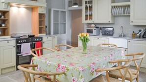 A kitchen with white cabinets, a stove, a microwave, and a dining table with a floral tablecloth. - Mayflower Apartment-UKC93 (Plymouth)