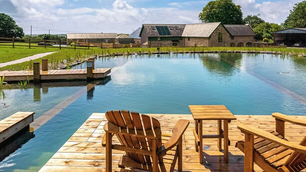 A wooden deck with chairs and tables by a calm pool, surrounded by buildings and greenery. - 3 bedroom accommodation in Kidwelly (Kidwelly)