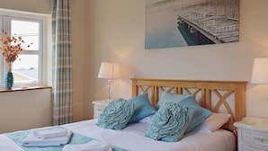 Un letto rifatto con un copriletto blu, cuscini bianchi e asciugamani piegati. Si vede una finestra con vista su un edificio e una tenda. Al muro, un quadro raffigura un molo.