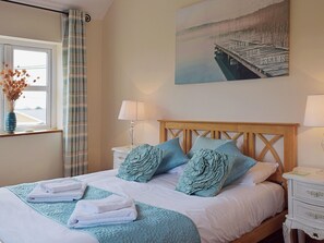 A neatly made bed with a blue quilt, white pillows, and folded towels. There is a window with a view of a building and a curtain. A painting of a dock is on the wall. - 2 bedroom accommodation in Kidwelly (Kidwelly)