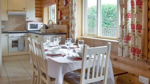 A wooden cabin with a kitchen and dining area. The kitchen has a microwave, sink, and stove. The dining area has a table set for four with chairs. There is a window with curtains and a view of greenery outside. - Pheasant Lodge-W43009 (Brayton, Selby)