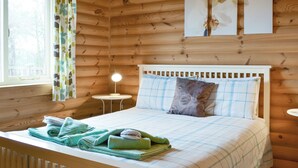 A wooden cabin bedroom with a bed, pillows, a bedside table with a lamp, and a window with curtains. - Pheasant Lodge-W43009 (Brayton, Selby)