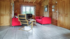 A log cabin interior with a wooden ceiling, a television, a red sofa, a wooden chair, and a window with curtains. - Pheasant Lodge-W43009 (Brayton, Selby)