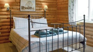 Una habitación de cabaña de madera con una cama, dos lámparas fijadas a la pared, un cuadro enmarcado y una ventana con cortinas.