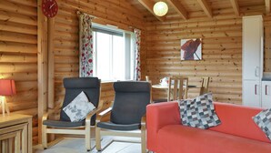 Interior de una cabaña de troncos con dos sillones, un sofá, una mesa de comedor y un reloj en la pared.