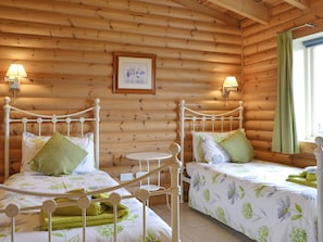 Una habitación tipo cabaña de madera con dos camas, cada una con estructura metálica y ropa de cama estampada con motivos florales. Hay lámparas fijadas a la pared y un cuadro enmarcado sobre las camas.