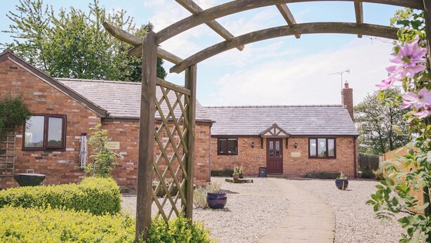 A brick cottage with a wooden archway entrance, a gravel driveway, and potted plants.