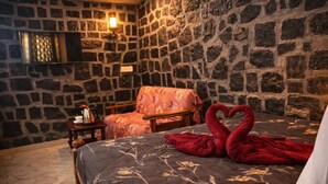 Una habitación acogedora con pared de piedra, cama con colcha estampada y una toalla roja con forma de cisne, además de una mesita con hervidor.
