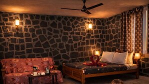 Una habitación acogedora con pared de piedra, cama de madera, sofá floral rojo y un niño sentado en el suelo.