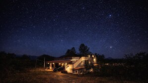 Una casa con una escalera y un coche estacionado frente a ella, rodeada por un cielo nocturno estrellado.