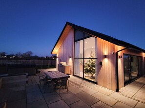 A modern wooden house with large glass windows, a patio with a table and chairs, and a visible Christmas tree inside. - Marsh Marigold Cottage - UKC4642 (Dunholme, near Lincoln)