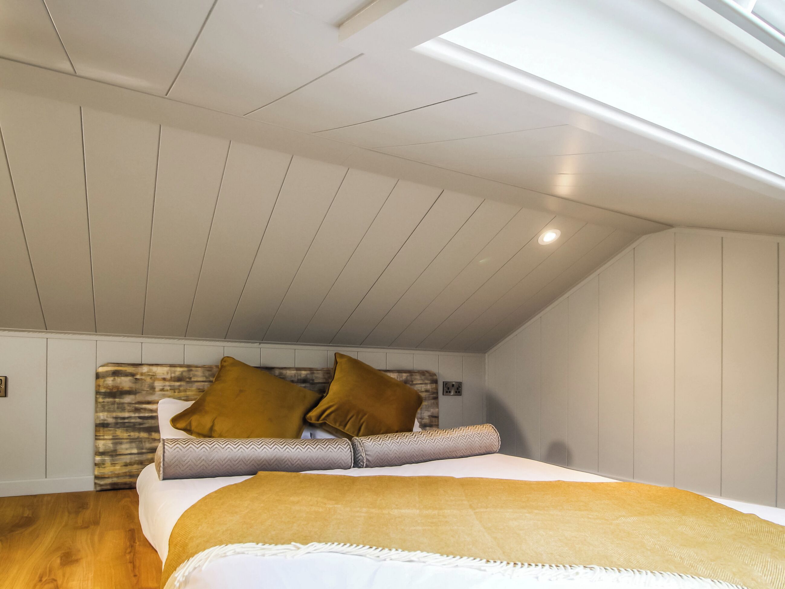 Quarto com teto inclinado, piso de madeira, cama com cobertor bege e duas almofadas.