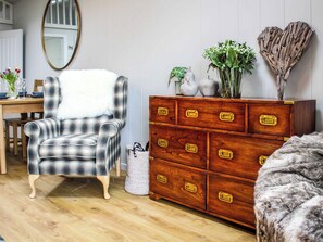 Ein Zimmer mit einem Karo-Sessel, einem Holzkleiderschrank mit Messinggriffen, einem Spiegel und einer Vase mit Blumen.