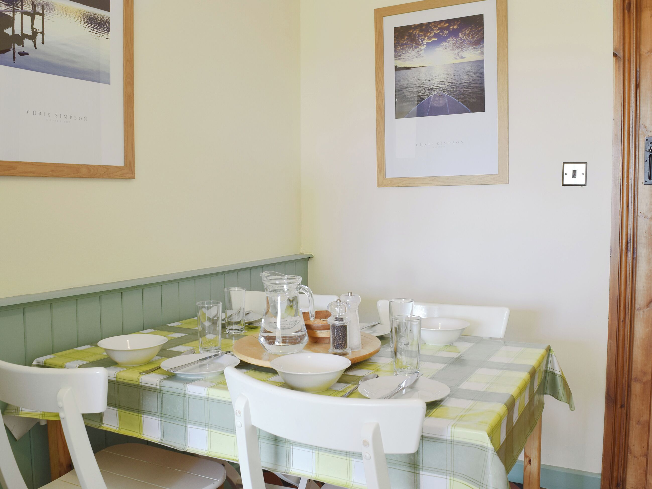 Mesa de comedor dispuesta con platos, vasos y una jarrón, rodeada de sillas blancas, con cuadros en las paredes.