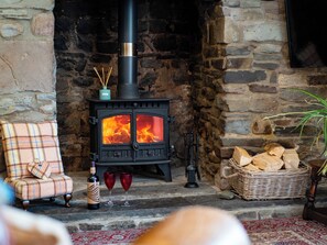 A stone fireplace with a black wood-burning stove, a plaid armchair, a bottle of wine, and a wicker basket of firewood. - 2 bedroom accommodation in Kidwelly (Kidwelly)