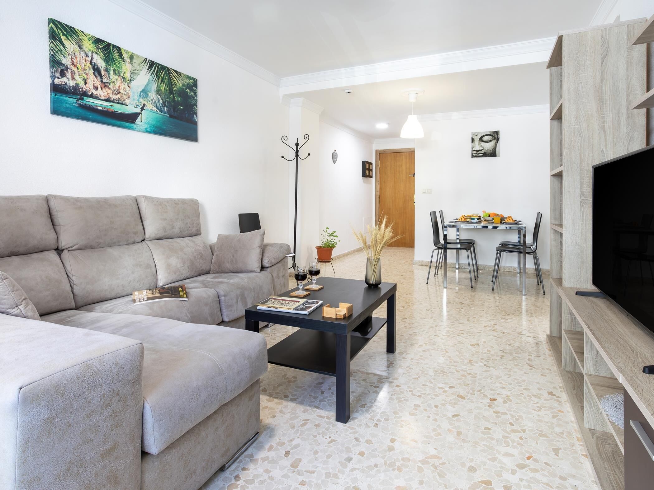 A modern living room with a grey sofa, a coffee table, and a TV. There is a dining area with a table and chairs in the background.