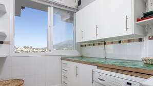 A modern kitchen with a washing machine, a window with a city view, and a wicker laundry basket. - Sunstay Adriana Flat II Fuengirola (Fuengirola)