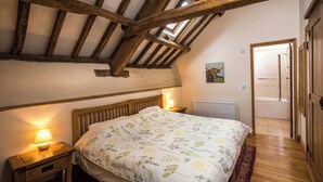 A bedroom with a wooden ceiling, a bed with floral bedding, a nightstand with a lamp, and a view of a bathroom through an open door. - The Old Stables - UKC4806 (Bishops Nympton)